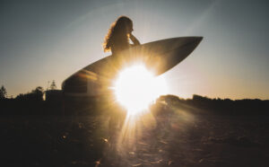 A picture of sun glinting through the silhouette of a woman carrying a surfboard