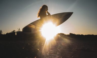 Still image from short film Ocean is She, of a woman carrying a surfboard and the setting sun glinting through underneath the board